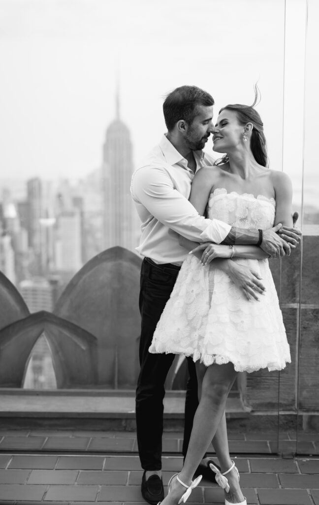 Black and white image of Felipe with his arms around Olya, her hair blowing softly in the wind at Top of the Rock.